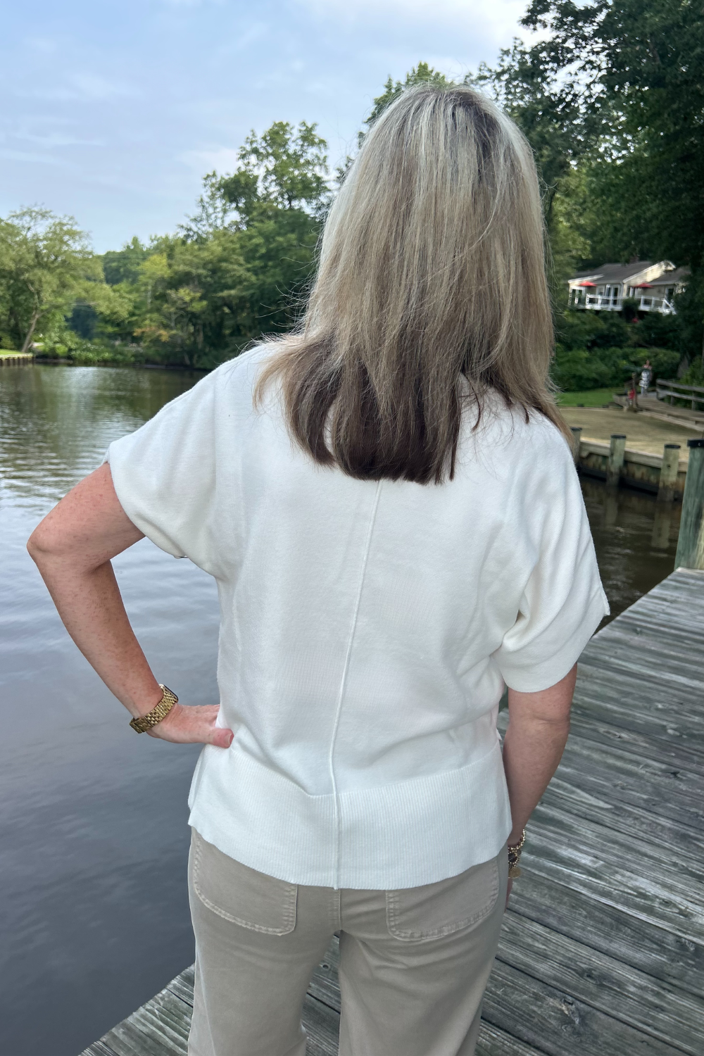A woman viewed from behind wearing a white short-sleeve top and beige pants, standing on a wooden dock by a lake with trees and a house in the background.