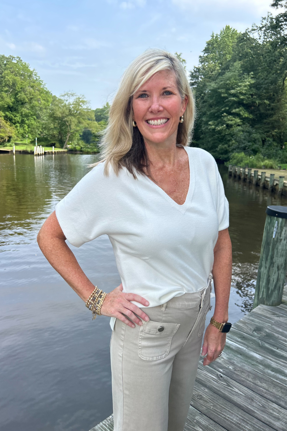 A smiling blonde woman stands on a wooden dock by a calm lake, wearing a white short-sleeve V-neck top and light beige pants.