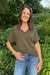 A woman wearing an olive green short-sleeve V-neck knit top and blue jeans, standing outdoors in front of a lush green background.
