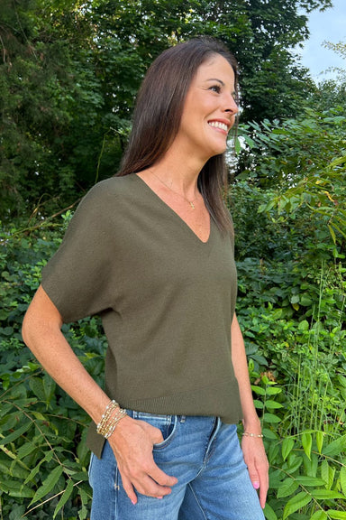 A woman wearing an olive green short-sleeve V-neck knit top and blue jeans, standing outdoors in front of a lush green background.