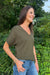 A woman wearing an olive green short-sleeve V-neck knit top and blue jeans, standing outdoors in front of a lush green background.