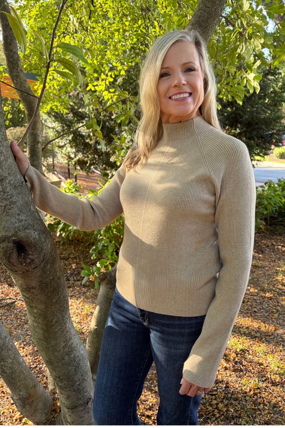 A smiling blonde woman wearing a beige ribbed mock neck sweater and dark blue jeans, standing outdoors next to a tree trunk in a sunny, leafy park setting.