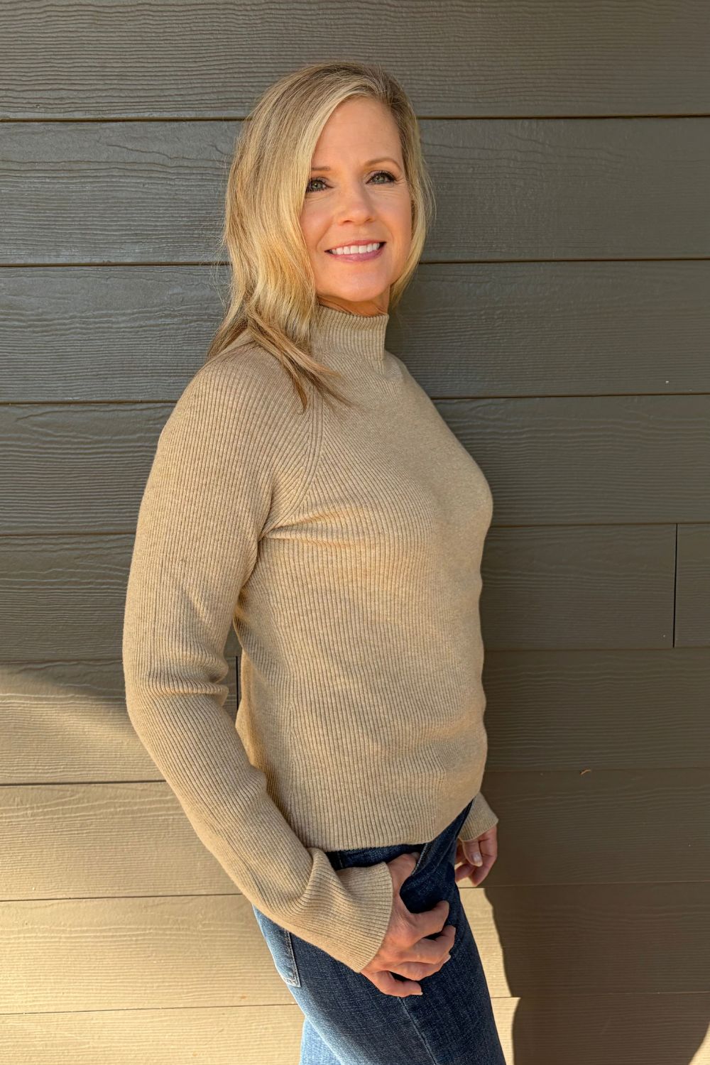 A smiling blonde woman wearing a beige ribbed mock-neck long-sleeve sweater and blue jeans, standing against a dark gray wood-paneled wall.