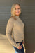 A smiling blonde woman wearing a beige ribbed mock-neck long-sleeve sweater and blue jeans, standing against a dark gray wood-paneled wall.