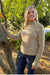 A smiling blonde woman wearing a beige ribbed mock neck sweater and dark blue jeans, standing outdoors next to a tree trunk in a sunny, leafy park setting.