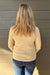 A woman viewed from behind wearing a beige ribbed knit sweater and blue jeans, standing against a gray wood-paneled wall.
