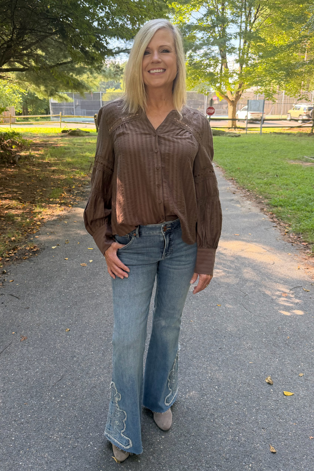 A woman standing on an outdoor paved path wearing a brown long-sleeve button-up blouse with textured vertical pleats and puffed shoulders, paired with light-wash blue flared jeans and beige shoes.