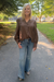 A woman standing on an outdoor paved path wearing a brown long-sleeve button-up blouse with textured vertical pleats and puffed shoulders, paired with light-wash blue flared jeans and beige shoes.
