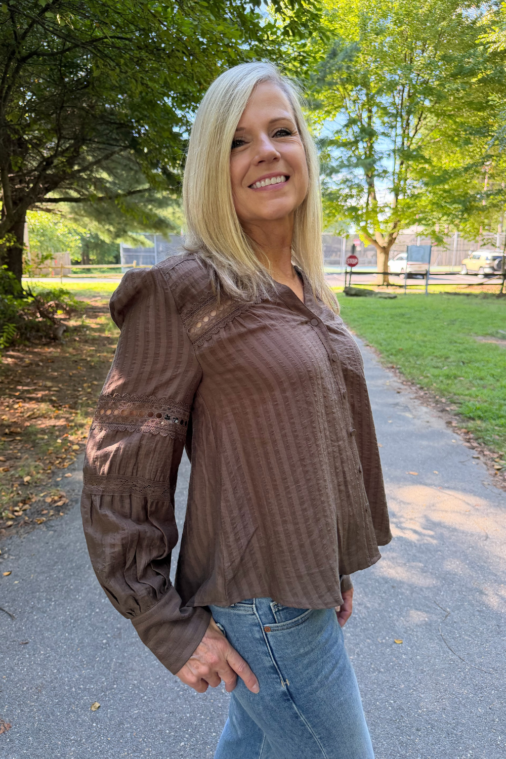 A smiling blonde woman wearing a brown textured long-sleeve blouse with lace detailing on the sleeves, paired with blue jeans, standing on an outdoor paved path surrounded by green trees.