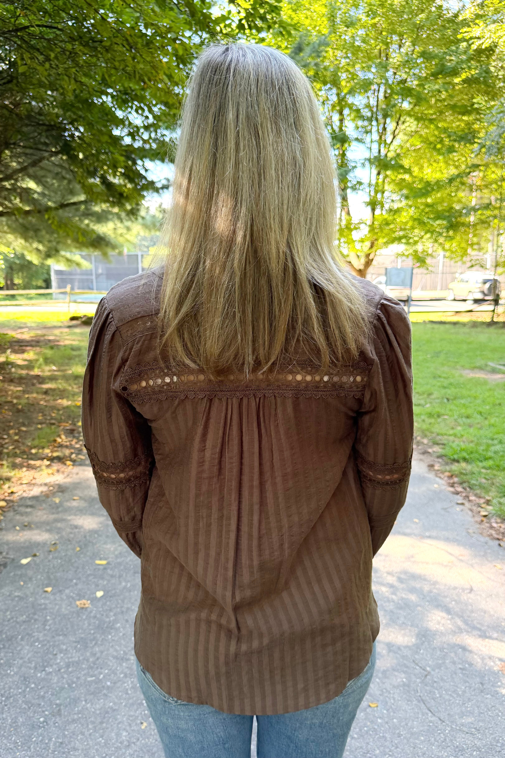 A woman viewed from behind wearing a brown long-sleeve blouse with vertical stripes and lace-like detailing across the upper back, standing on an outdoor paved path with trees and grass in the background.