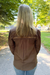 A woman viewed from behind wearing a brown long-sleeve blouse with vertical stripes and lace-like detailing across the upper back, standing on an outdoor paved path with trees and grass in the background.