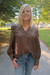 A woman with blonde hair wearing a brown long-sleeve button-up blouse with vertical stripes and lace-up front details, paired with blue jeans, standing outdoors on a paved path in a park.