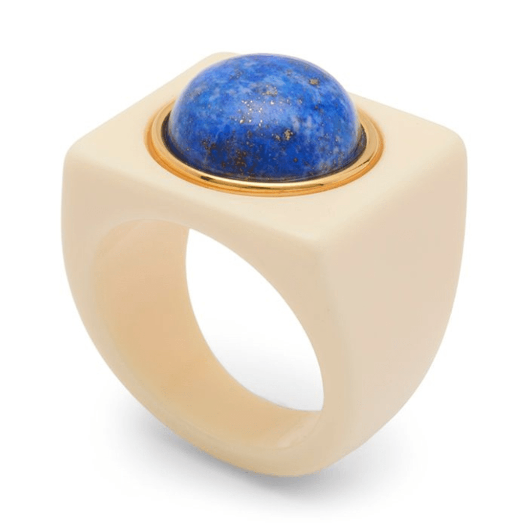 A statement ring featuring a large, matte cream-colored square bezel with a polished blue lapis lazuli sphere set in a gold-tone bezel, shown on a white background.