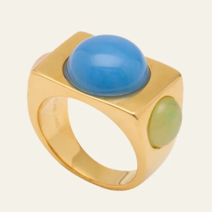 A gold signet-style ring featuring a large, smooth oval blue cabochon on the top and a smaller oval light green cabochon on the side, set against a solid off-white background.