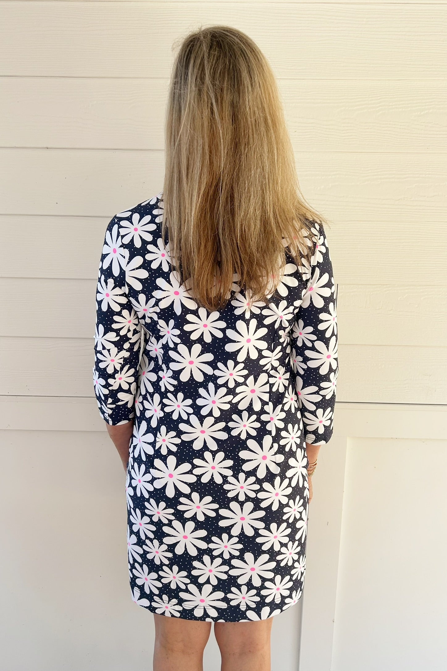 A woman viewed from behind wearing a navy blue long-sleeve dress with a white daisy pattern that has pink centers, standing against a white paneled wall.