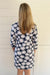 A woman viewed from behind wearing a navy blue long-sleeve dress with a white daisy pattern that has pink centers, standing against a white paneled wall.