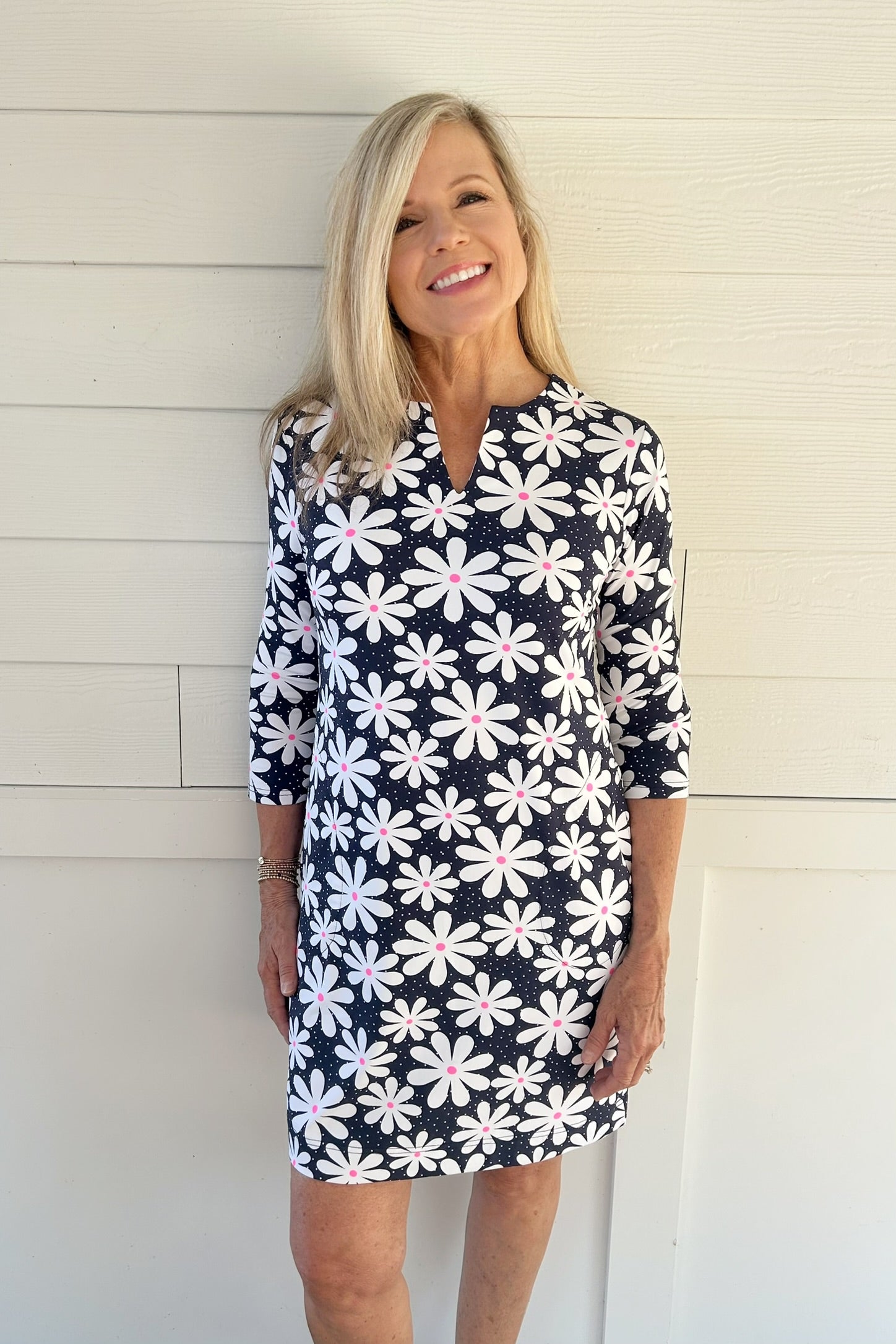 A smiling blonde woman wearing a navy blue and white floral print mini dress with three-quarter sleeves, standing against a white paneled wall.