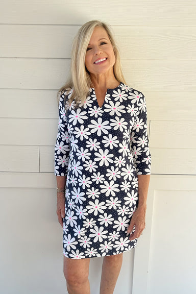 A smiling blonde woman wearing a navy blue and white floral print mini dress with three-quarter sleeves, standing against a white paneled wall.
