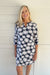 A smiling blonde woman wearing a navy blue and white floral print mini dress with three-quarter sleeves, standing against a white paneled wall.