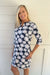 A smiling blonde woman wearing a navy blue knee-length dress with a white and pink daisy floral pattern, standing against a white wood-paneled wall.