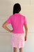 A woman shown from the back wearing a bright pink short-sleeve polo shirt and a white golf skirt with a pink seashell and starfish pattern, standing against a white paneled wall.