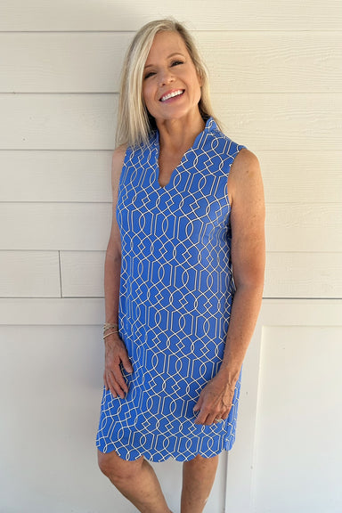 A smiling blonde woman wearing a sleeveless blue dress with a white geometric pattern, standing against a white paneled wall.
