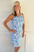 A smiling blonde woman wearing a sleeveless bodycon dress with a blue, teal, and white abstract pattern, standing against a white paneled wall.