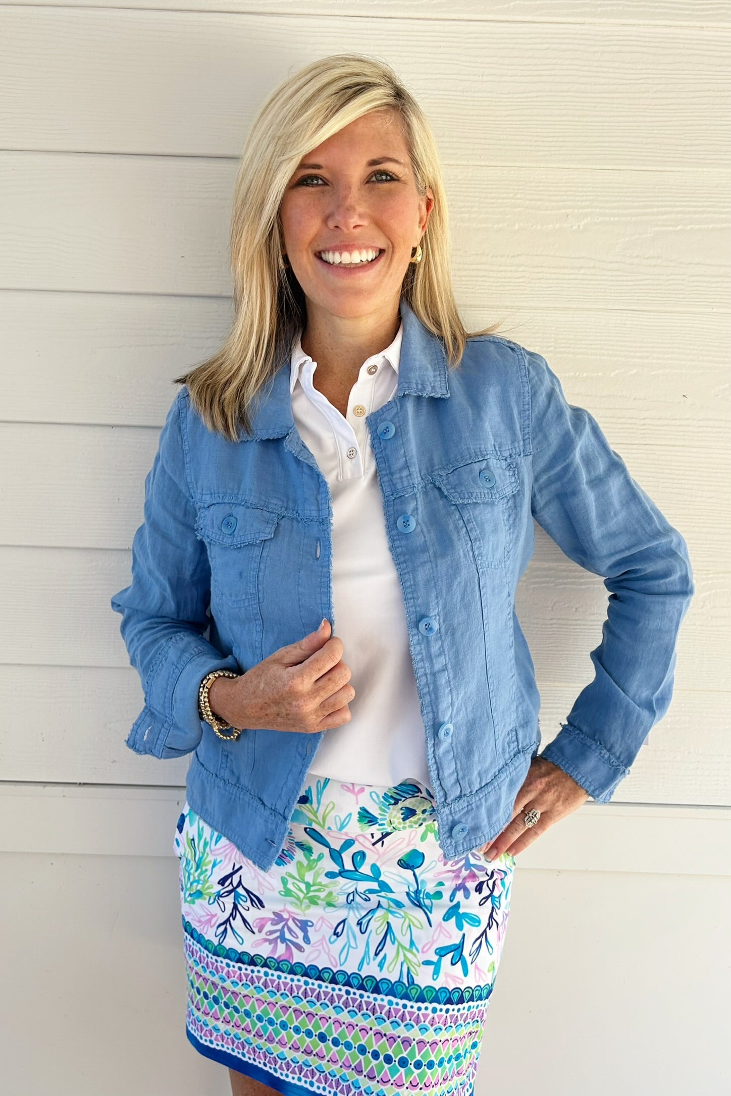 A smiling blonde woman stands against a white paneled wall wearing a light blue denim jacket over a white collared shirt and a colorful floral and geometric patterned skirt.