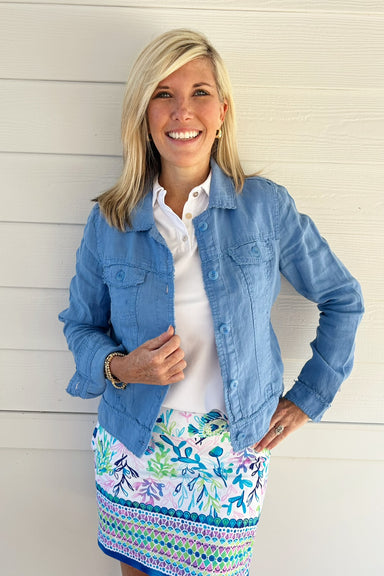 A smiling blonde woman stands against a white paneled wall wearing a light blue denim jacket over a white collared shirt and a colorful floral and geometric patterned skirt.