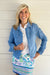 A smiling blonde woman stands against a white paneled wall wearing a light blue denim jacket over a white collared shirt and a colorful floral and geometric patterned skirt.