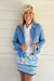 A smiling blonde woman stands against a white paneled wall wearing a light blue denim jacket over a white collared shirt and a white mini skirt with a colorful blue, green, and purple floral and geometric pattern.