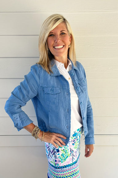 A smiling blonde woman wearing a light blue denim jacket over a white collared shirt and a colorful floral patterned skirt, standing against a white wood-paneled wall.