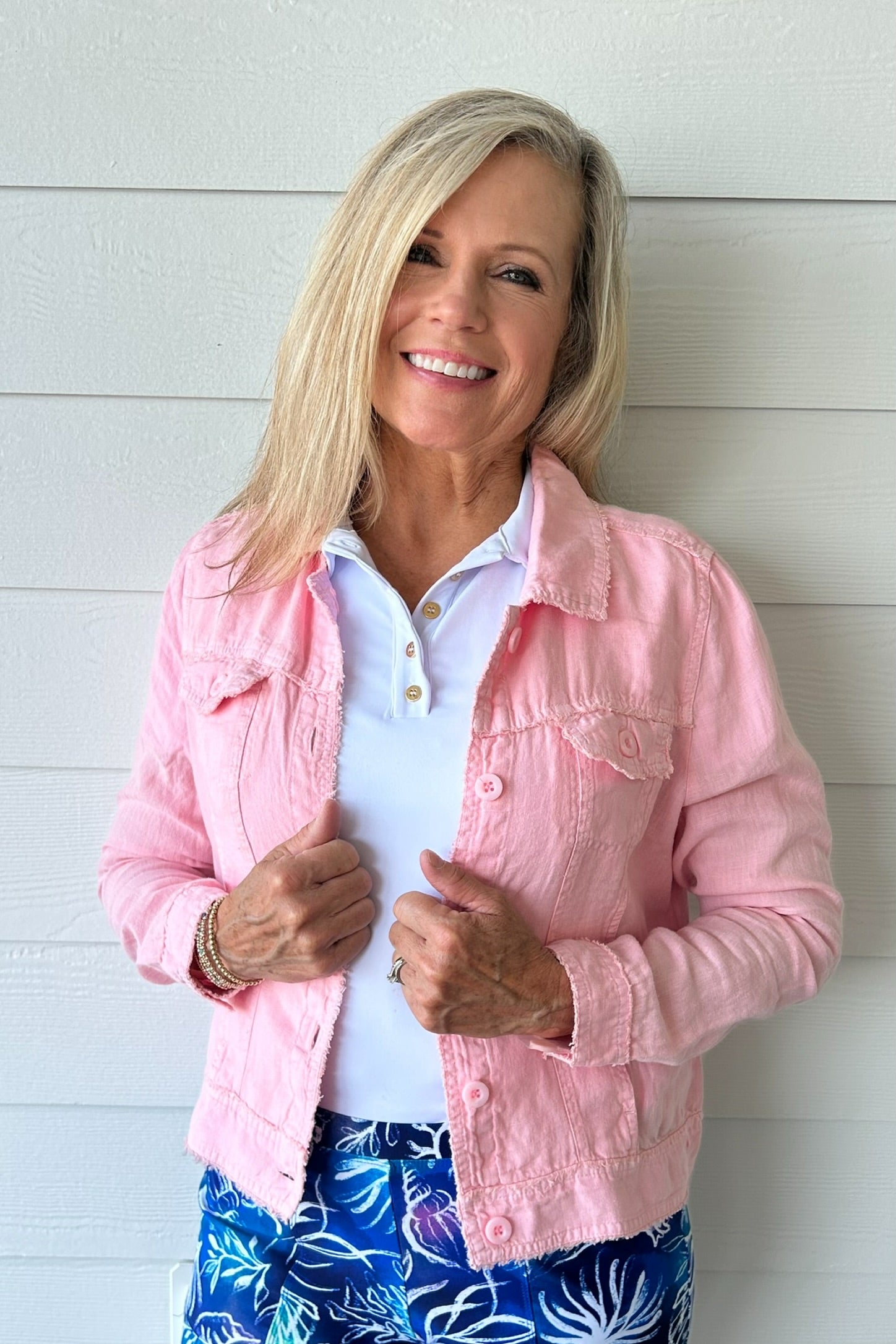 A smiling blonde woman wearing a light pink textured denim jacket over a white polo shirt and blue floral pants, standing against a white shiplap wall.