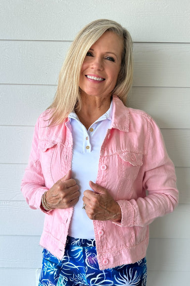 A smiling blonde woman wearing a light pink textured denim jacket over a white polo shirt and blue floral pants, standing against a white shiplap wall.