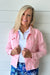 A smiling blonde woman wearing a light pink textured denim jacket over a white polo shirt and blue floral pants, standing against a white shiplap wall.