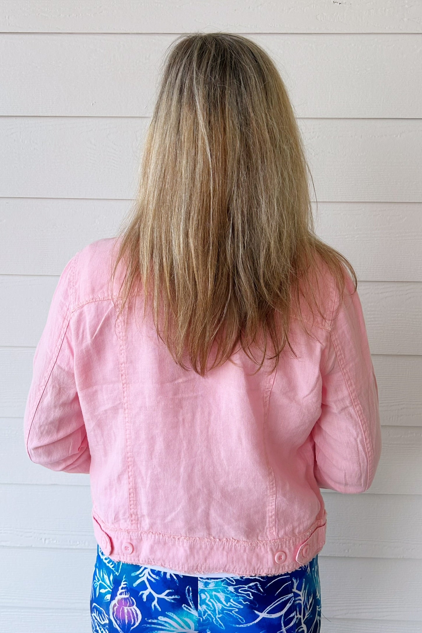 A woman viewed from behind wearing a light pink cropped jacket and blue floral pants, standing against a white shiplap wall.