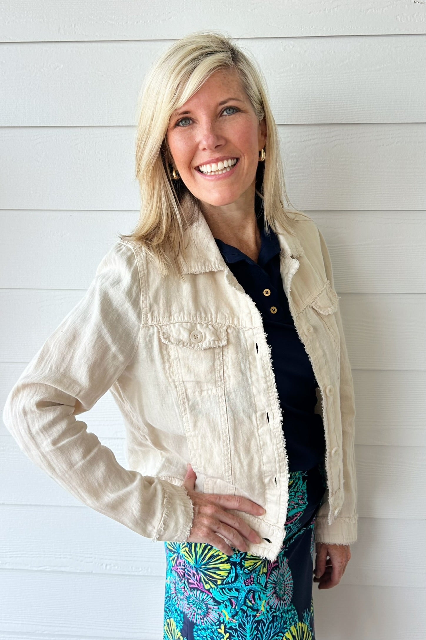 A smiling blonde woman wearing a light beige linen-textured cropped jacket over a navy top and a colorful floral skirt, standing against a white shiplap wall.