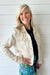 A smiling blonde woman wearing a light beige linen-textured cropped jacket over a navy top and a colorful floral skirt, standing against a white shiplap wall.