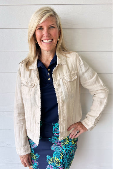 A smiling blonde woman stands against a white shiplap wall wearing a light beige linen jacket over a navy polo shirt and a navy skirt with a colorful tropical floral print.