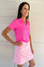 A woman modeling a bright pink short-sleeve polo shirt and a pink and white patterned skirt, standing against a white paneled wall.