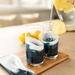 Two clear glasses filled with a dark blue drink and lemon slices, set on a wooden tray with a glass pitcher pouring water in a bright indoor setting.