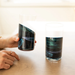 Two clear drinking glasses with a dark marbled pattern in black, teal, and blue, shown on a wooden table with a hand holding one glass.