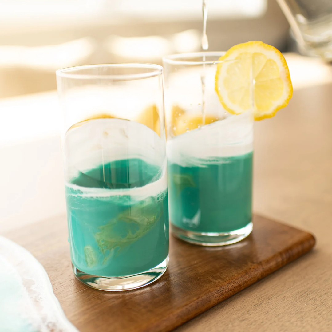 Two tall clear glasses filled with bright turquoise liquid, garnished with lemon slices, sitting on a wooden board in a warm indoor setting.