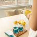 A person pours clear liquid from a glass pitcher into two tall glasses containing bright blue liquid, with lemon slices visible in the pitcher and lemons on the table. The scene is set on a light wood table near a window in a bright indoor environment.