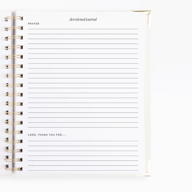 A white spiral-bound devotional journal with lined pages, featuring gold-toned metal spiral binding and printed text including 'PRAYER', 'devotional journal', and 'LORD, THANK YOU FOR.