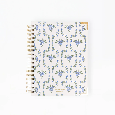 A spiral-bound devotional journal with a white cover decorated in a repeating pattern of blue flowers and green leaves, featuring gold-toned metal spiral binding and a small gold accent tab on the top right.