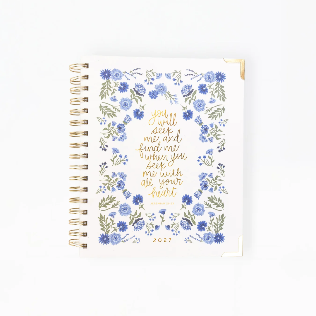 A white 2027 spiral-bound planner with a gold wire binding and a central gold quote surrounded by blue and green floral illustrations, shown on a clean white background.