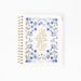 A white 2027 spiral-bound planner with a gold wire binding and a central gold quote surrounded by blue and green floral illustrations, shown on a clean white background.