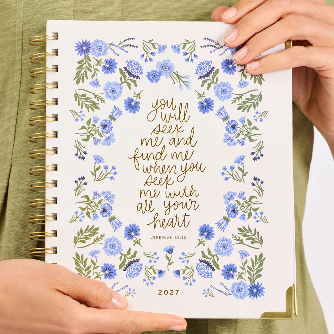 A person holds a 2027 spiral-bound journal featuring a white cover decorated with blue flowers and green leaves, with gold foil lettering that reads 'you will seek me and find me when you seek me with all your heart' and 'JERUSALEM 29:13'.