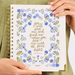 A person holds a 2027 spiral-bound journal featuring a white cover decorated with blue flowers and green leaves, with gold foil lettering that reads 'you will seek me and find me when you seek me with all your heart' and 'JERUSALEM 29:13'.